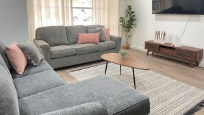 Un salon avec un canapé gris, une table basse, un meuble TV en bois et une grande fenêtre avec des rideaux.