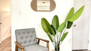 Un fauteuil gris, recouvert de tissu, avec des pieds en bois, une plante en pot aux grandes feuilles vertes et une porte blanche dans une pièce dont le sol est recouvert de planches de bois de couleur claire.