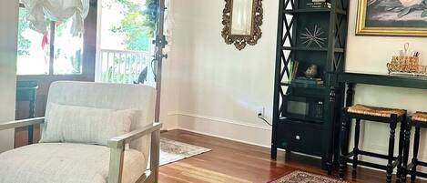 A living room with a wooden floor, a white armchair, a black shelving unit, a red patterned rug, and a framed mirror on the wall.
