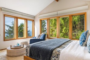 Une chambre dotée d’un grand lit, d’une chaise, d’une table ronde et de plusieurs fenêtres donnant sur des arbres et une étendue d’eau.