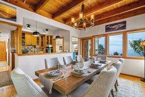 Un coin repas avec une table en bois dressée pour six personnes, une cuisine avec des placards en bois et de grandes fenêtres offrant une vue dégagée.