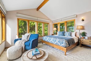 Ein Schlafzimmer mit einem großen Bett, einem blauen Sessel, einem runden Couchtisch mit Büchern und einem Nachttisch aus Holz.