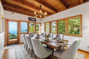 Comedor con mesa de madera dispuesta para seis personas, ventanas amplias con vistas a follaje y una araña.