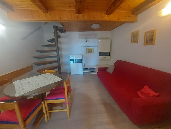 Une pièce chaleureuse avec un canapé rouge, une table à manger en bois entourée de chaises, un petit coin cuisine équipé d’un réfrigérateur et d’un four à micro-ondes, et un escalier menant à un étage supérieur.