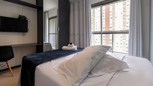 Habitación de hotel con una cama bien hecha, un escritorio con silla y un ventanal con vistas a edificios altos.