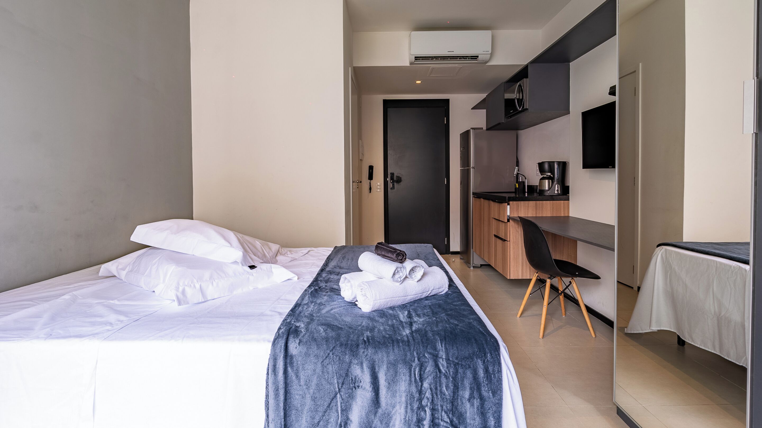 A hotel room with a bed, a desk, a chair, and a kitchenette.