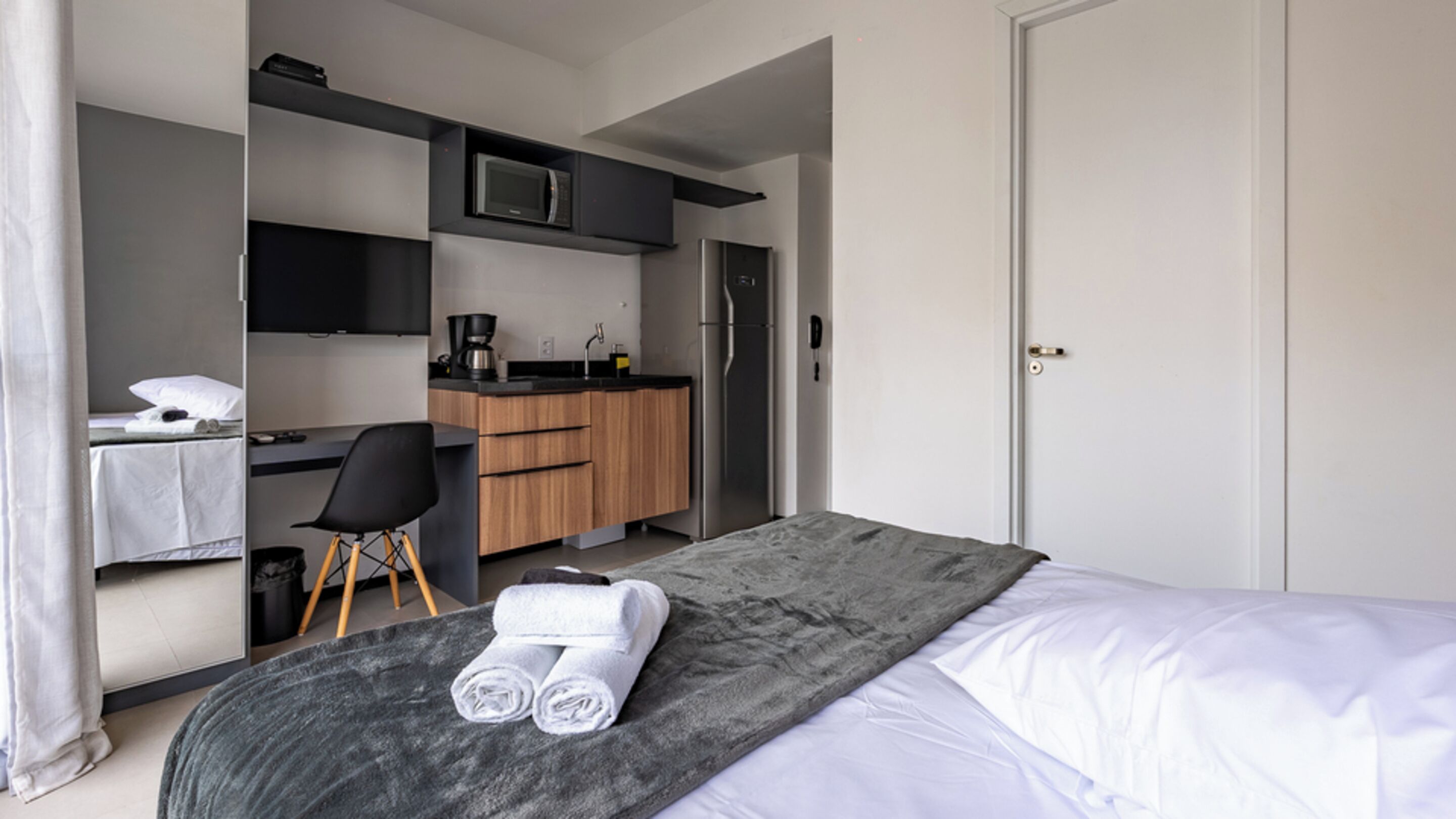 A hotel room with a bed, a desk, a chair, a microwave, a refrigerator, and a small kitchenette.