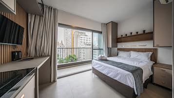 A modern hotel room with a bed, bedside table, TV, and a balcony with a city view.
