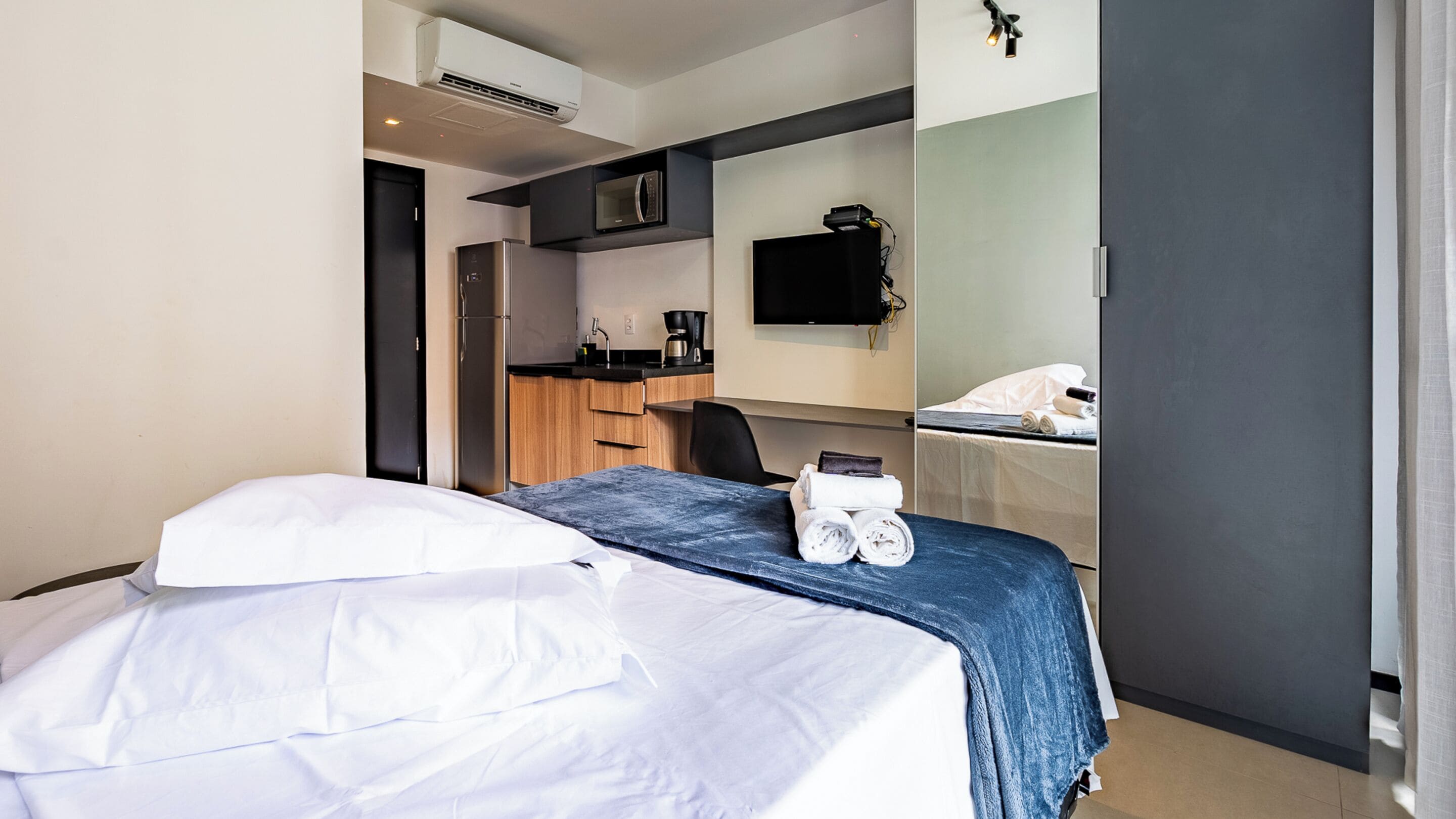 Quarto de hotel com cama, escrivaninha, micro-ondas, cafeteira e televisão.