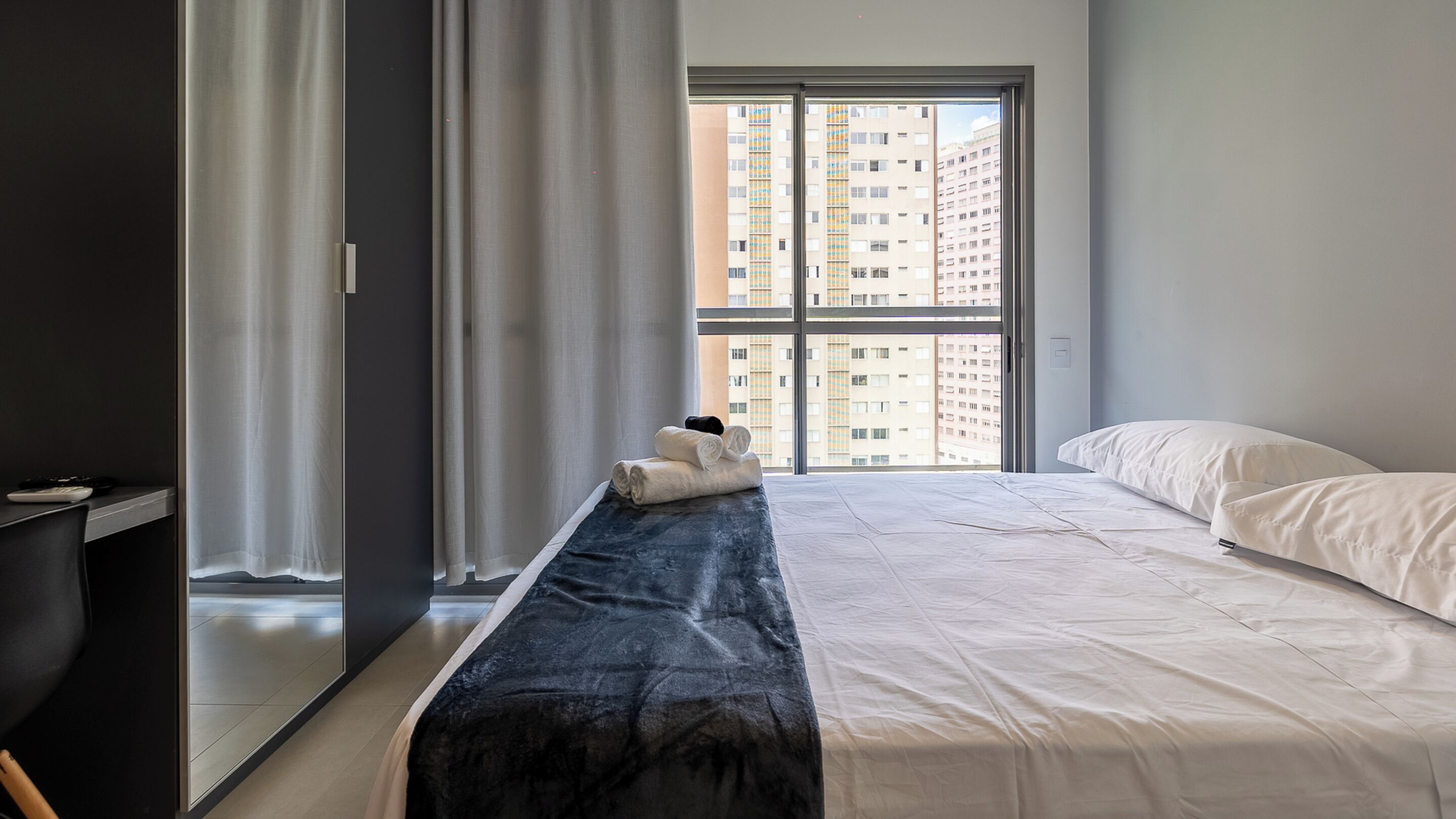 A modern hotel room with a large bed, a bench with towels, a desk, and a view of high-rise buildings through a large window.