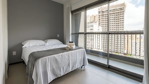 Un dormitorio moderno con un ventanal grande, una cama con ropa de cama blanca y vistas a rascacielos.