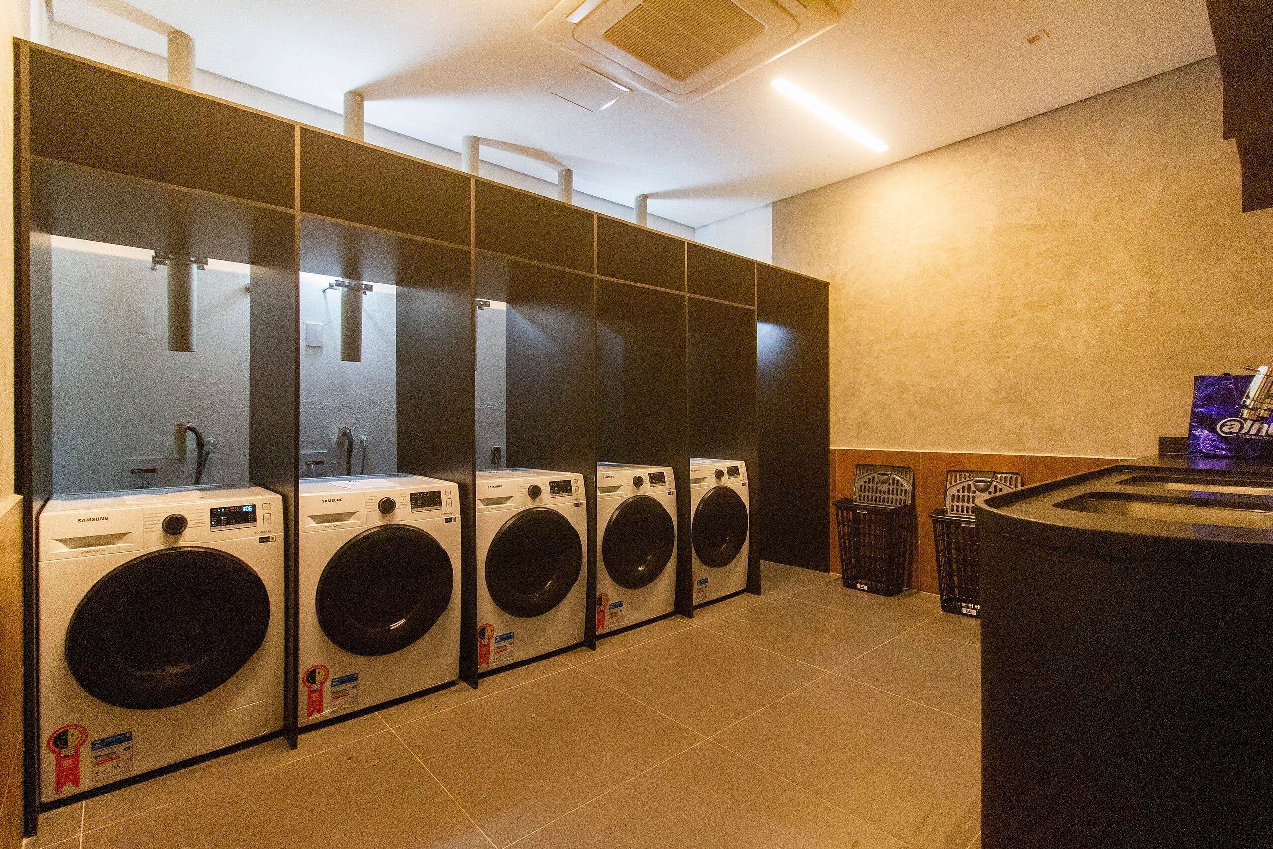 A row of Samsung front-loading washing machines in a public laundry area with a trash bin and recycling bins nearby.