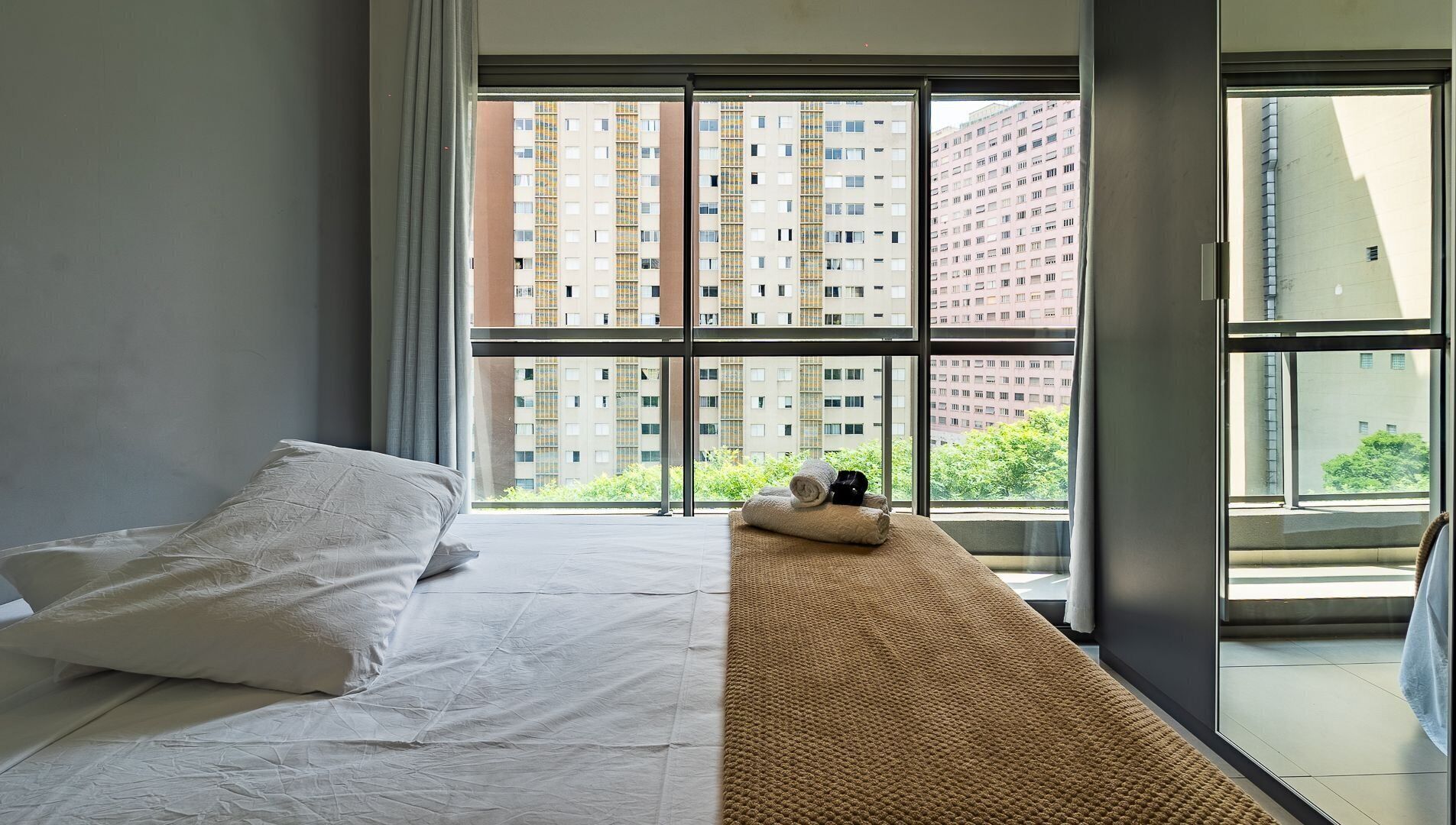 Un dormitorio con un ventanal grande, una cama y vistas a edificios altos.