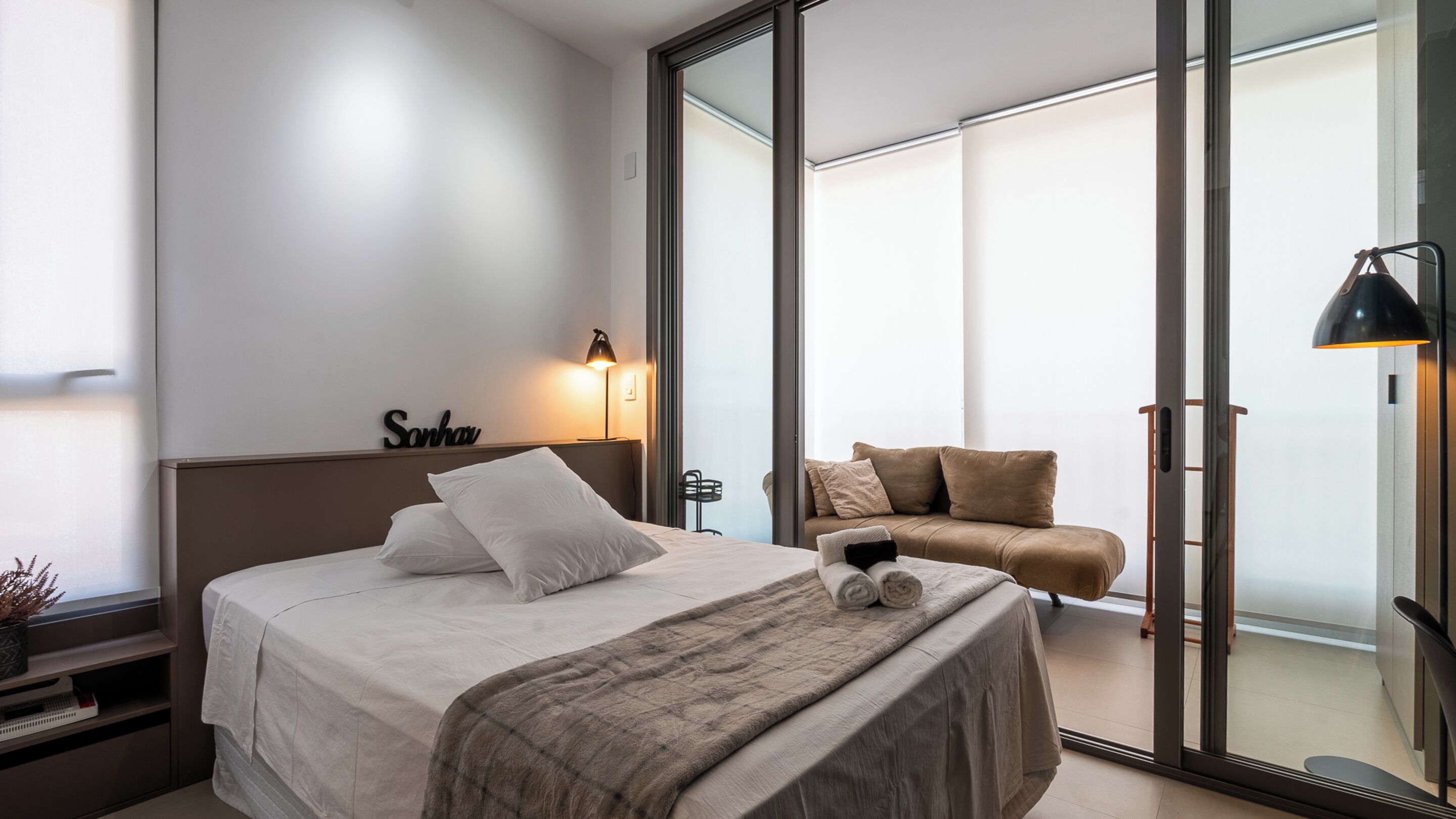 Een moderne slaapkamer met een groot bed, een bank, een bureau en een glazen schuifdeur die leidt naar een balkon.
