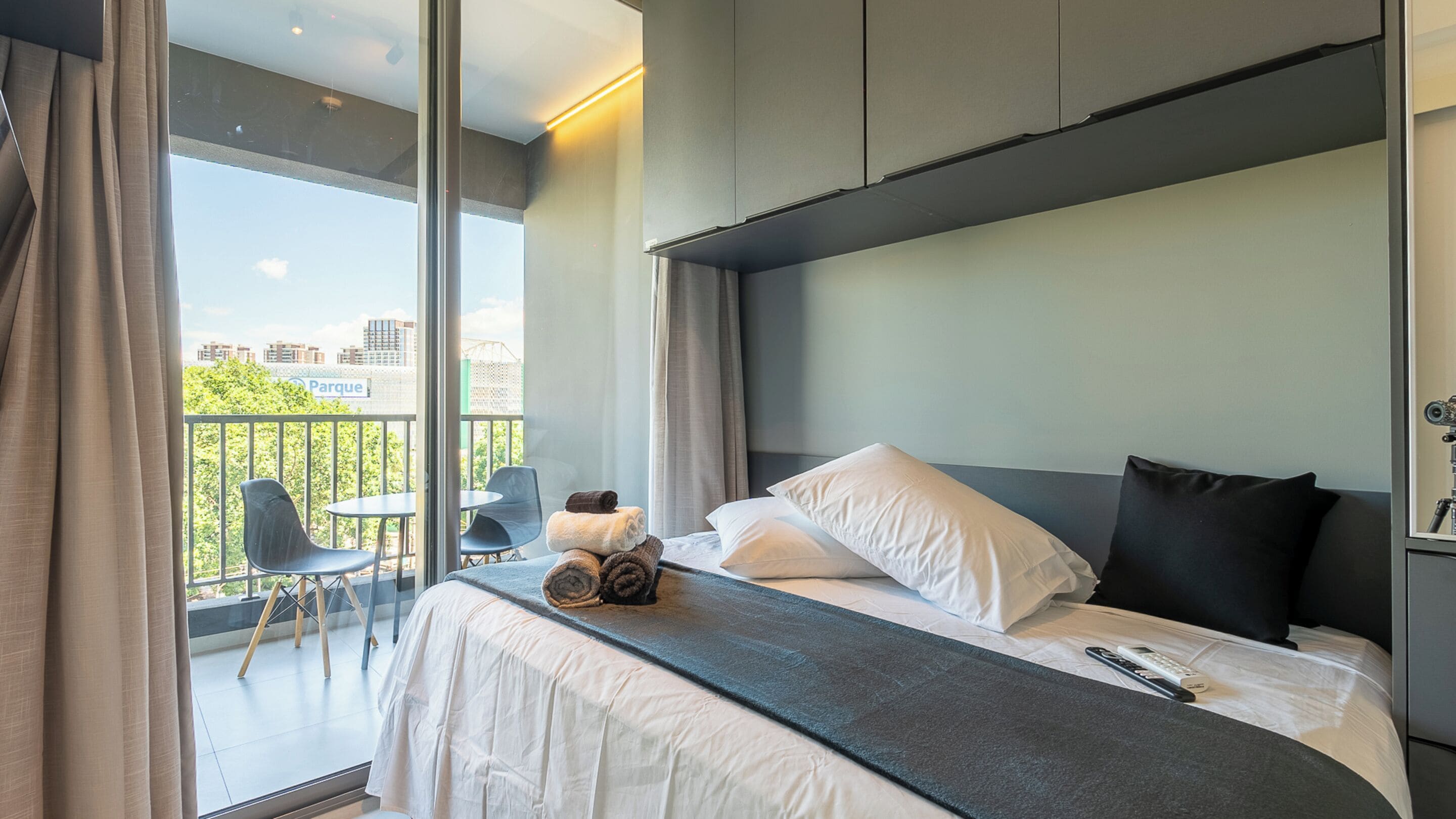 A modern hotel room with a large bed, a balcony with chairs, and a view of a park.