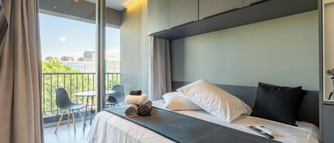 A modern hotel room with a large bed, a balcony with chairs, and a view of a park.