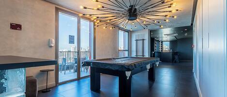 Una mesa de billar en una habitación amplia con una lámpara de techo moderna y ventanas grandes.