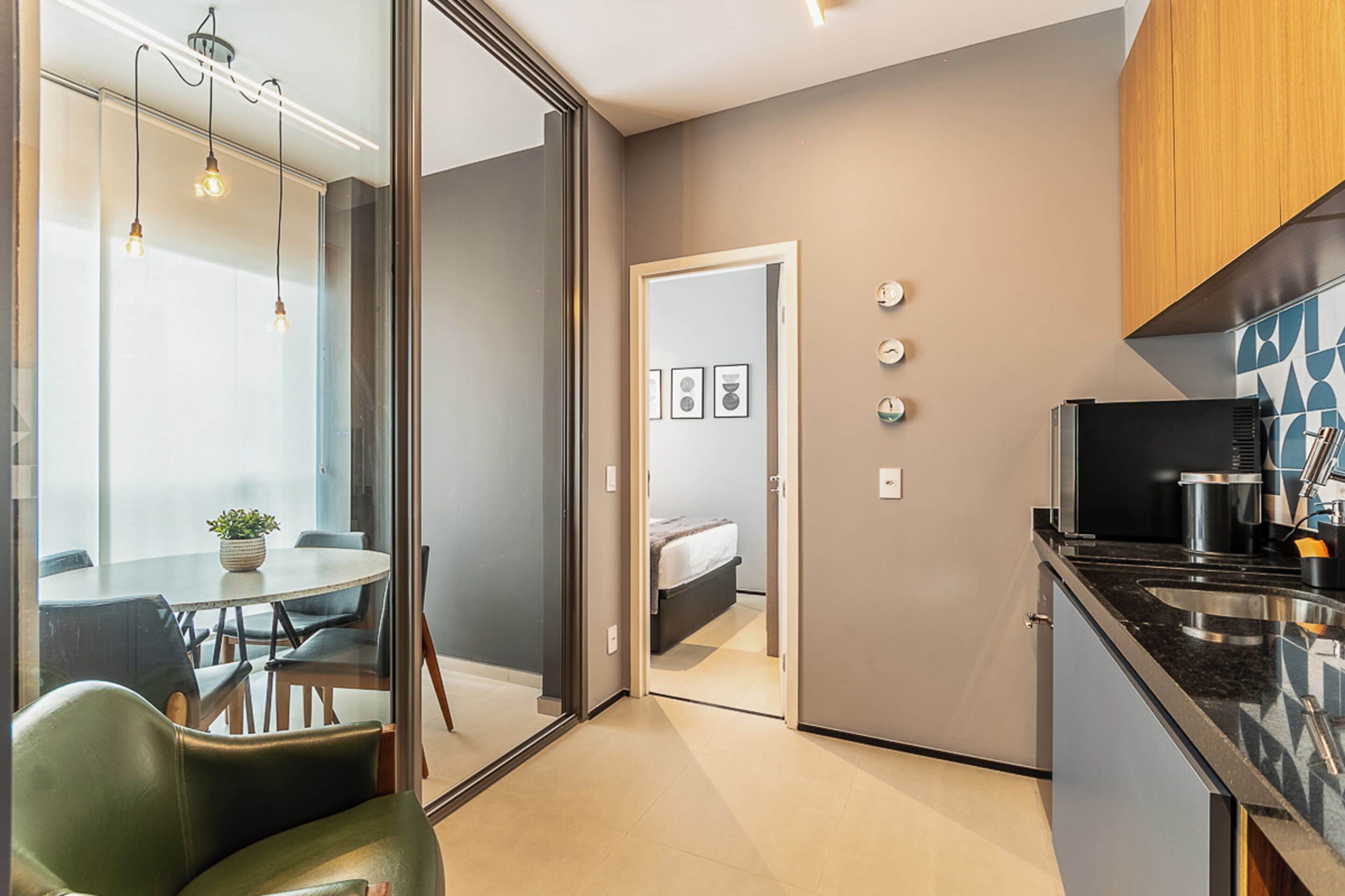A modern kitchen with a microwave, stove, and sink. There is a dining area with a table and chairs, and a glass door leading to a bedroom.
