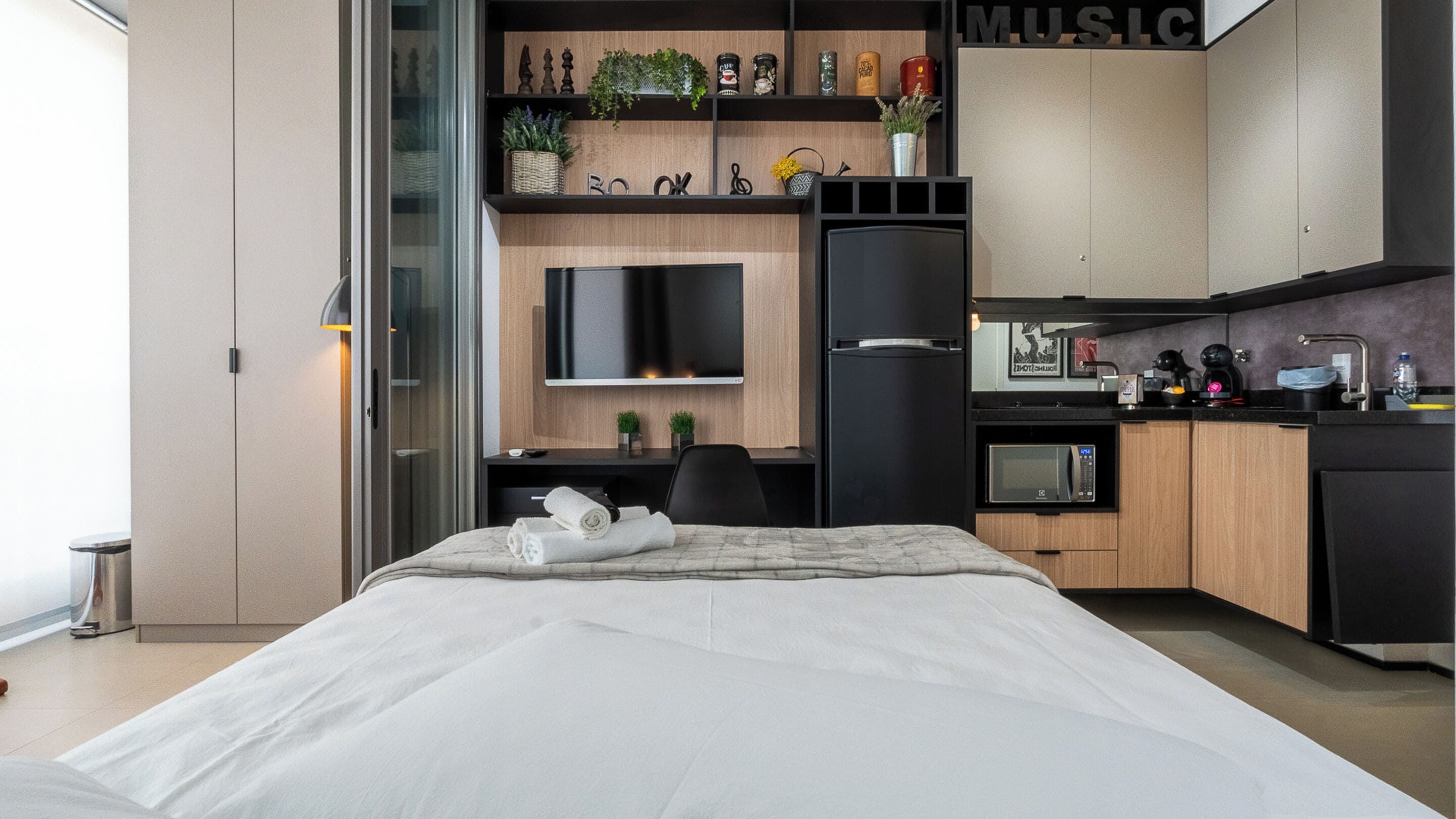 A modern bedroom with a bed, a desk, a chair, a TV, and a kitchenette with a microwave and coffee maker.