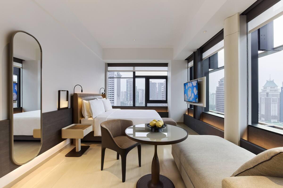 A modern hotel room with a bed, a seating area with a table, a TV, and large windows with a city view.