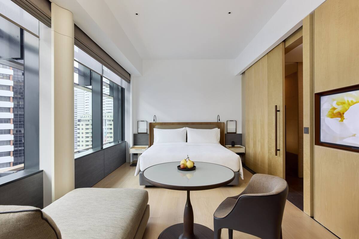 A modern hotel room with a large bed, a round table with fruit, a sofa, and a TV on the wall.