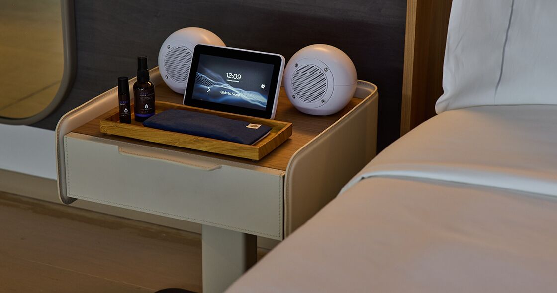 Una mesita de noche con una tableta, dos altavoces y una botella de loción en una habitación de hotel.