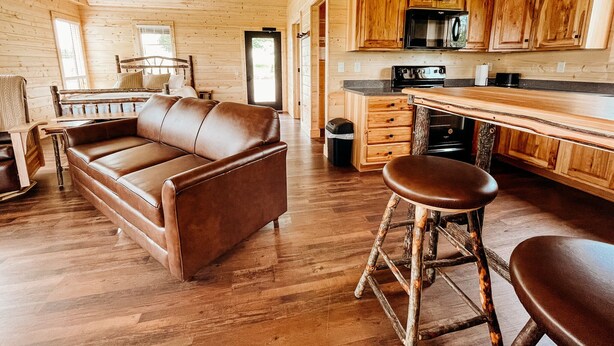 A cozy cabin interior with a wooden floor, a leather sofa, a wooden dining table with stools, and a kitchen area with wooden cabinets and appliances. - The Elk Cabin - Hidden Acres Cabins (Bethany)