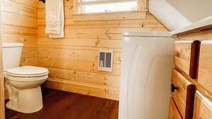 A compact wooden cabin bathroom with a toilet, a window with blinds, and a white refrigerator. - The Elk Cabin - Hidden Acres Cabins (Bethany)