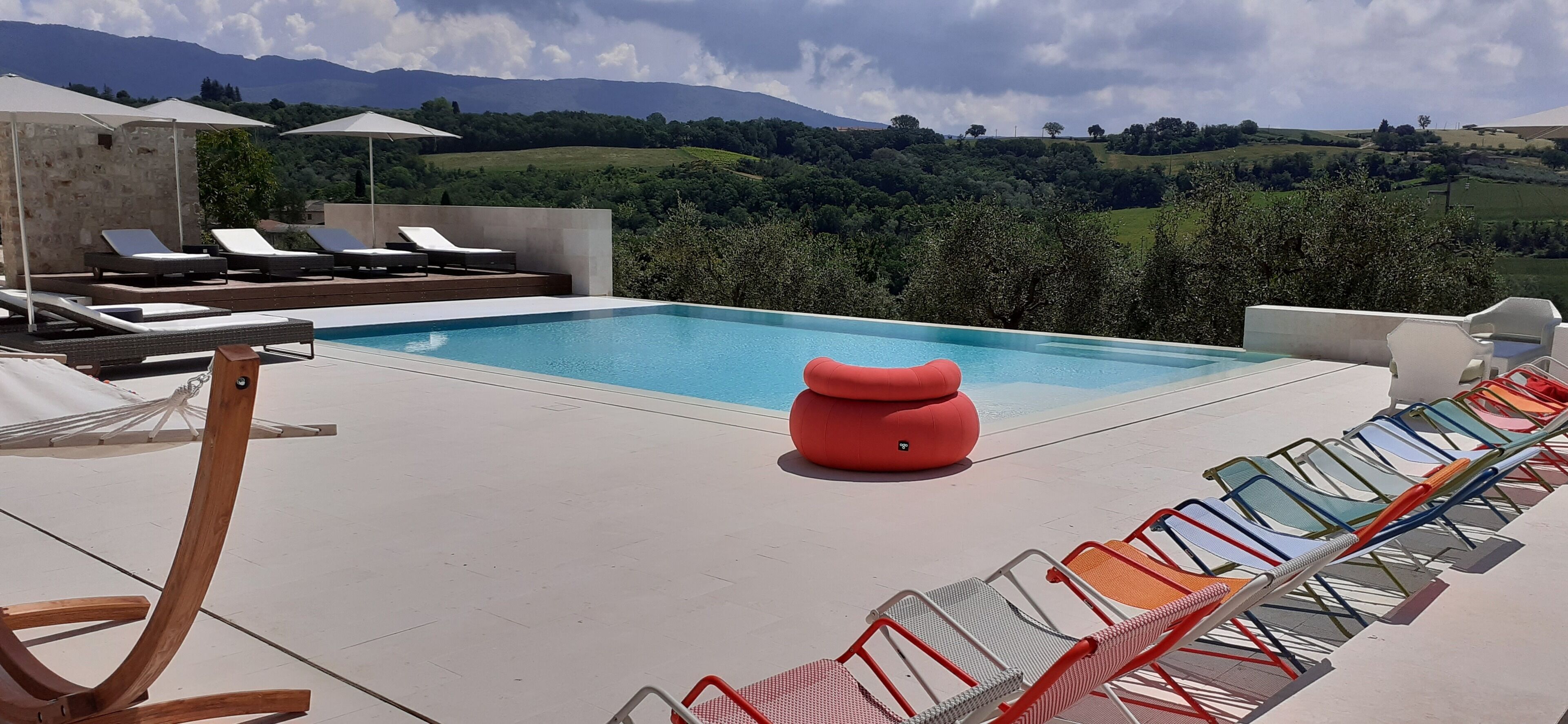 A pool area with lounge chairs and a red ottoman, surrounded by a scenic view of hills and trees.