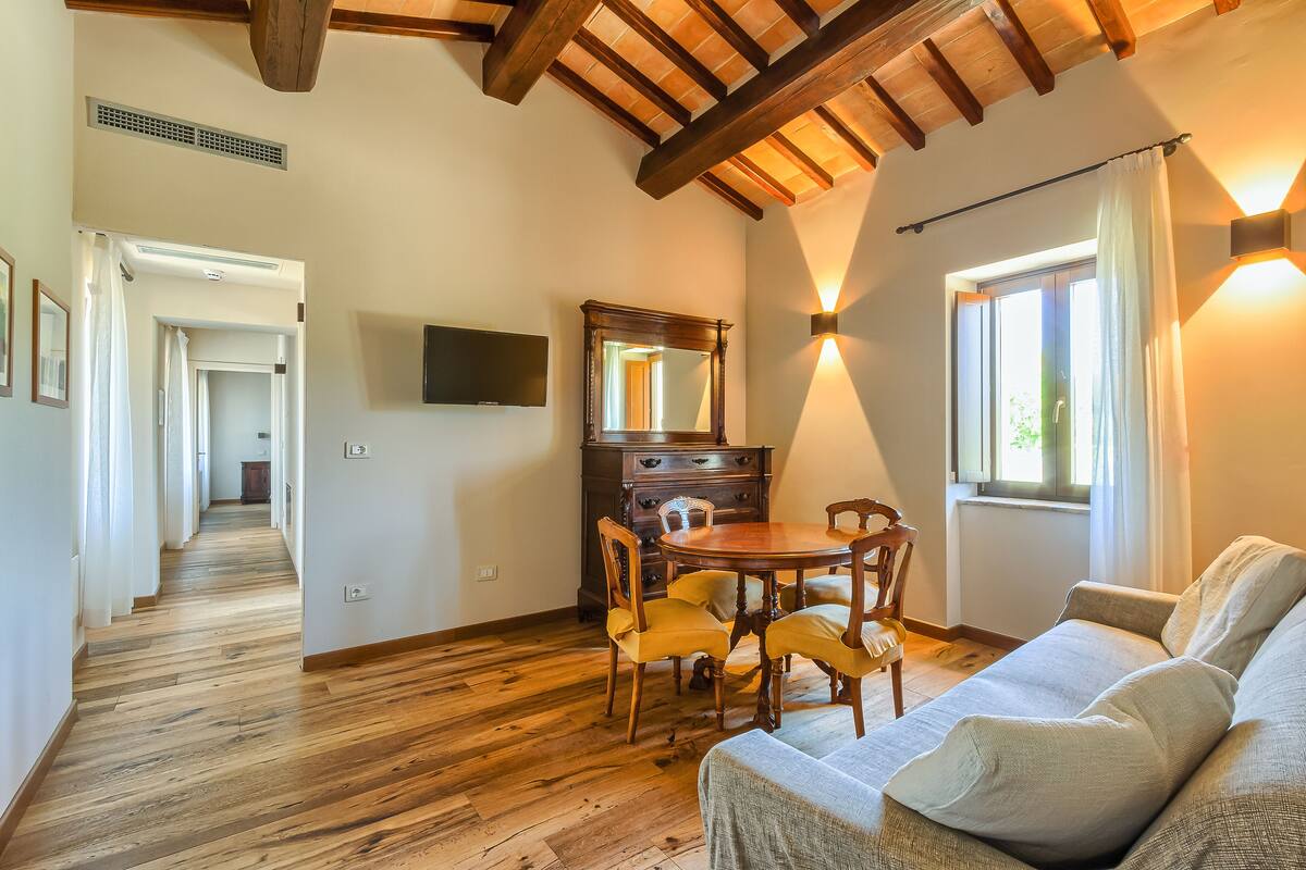 Una habitación con suelo de madera, una mesa de comedor con sillas, un sofá, un espejo grande y una ventana con cortinas.