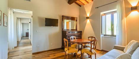 A room with a wooden floor, a dining table with chairs, a sofa, a large mirror, and a window with curtains.
