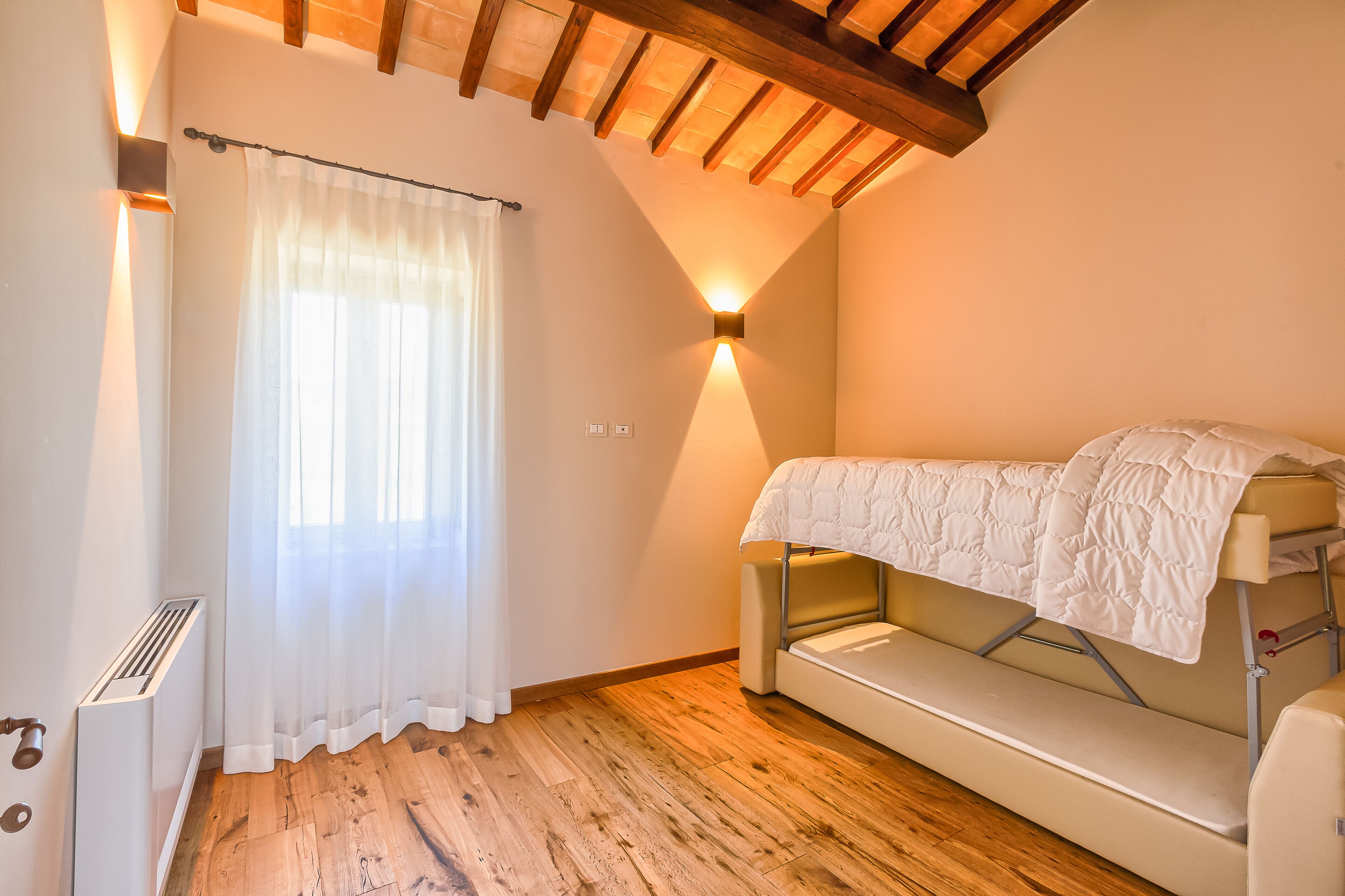 A small, cozy room with a bunk bed, wooden flooring, a radiator, and a window with sheer curtains.