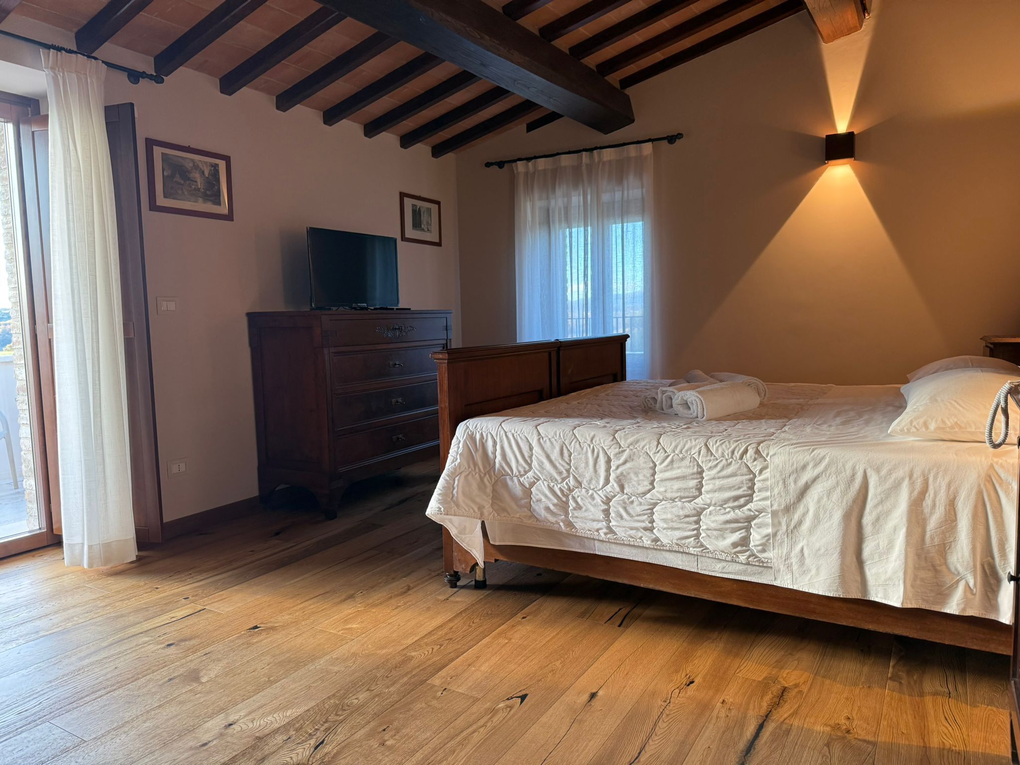 A bedroom with a wooden bed, a dresser, a television on top, and a window with sheer curtains.