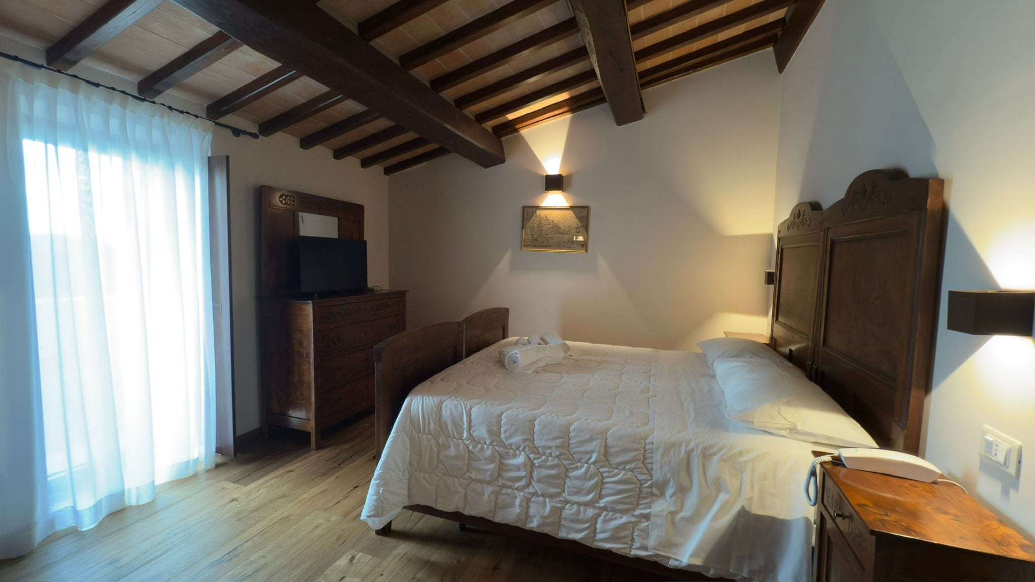 Een slaapkamer met een houten plafond, een bed met een witte deken, een houten dressoir, een bureau met een lamp en een raam met dunne gordijnen.