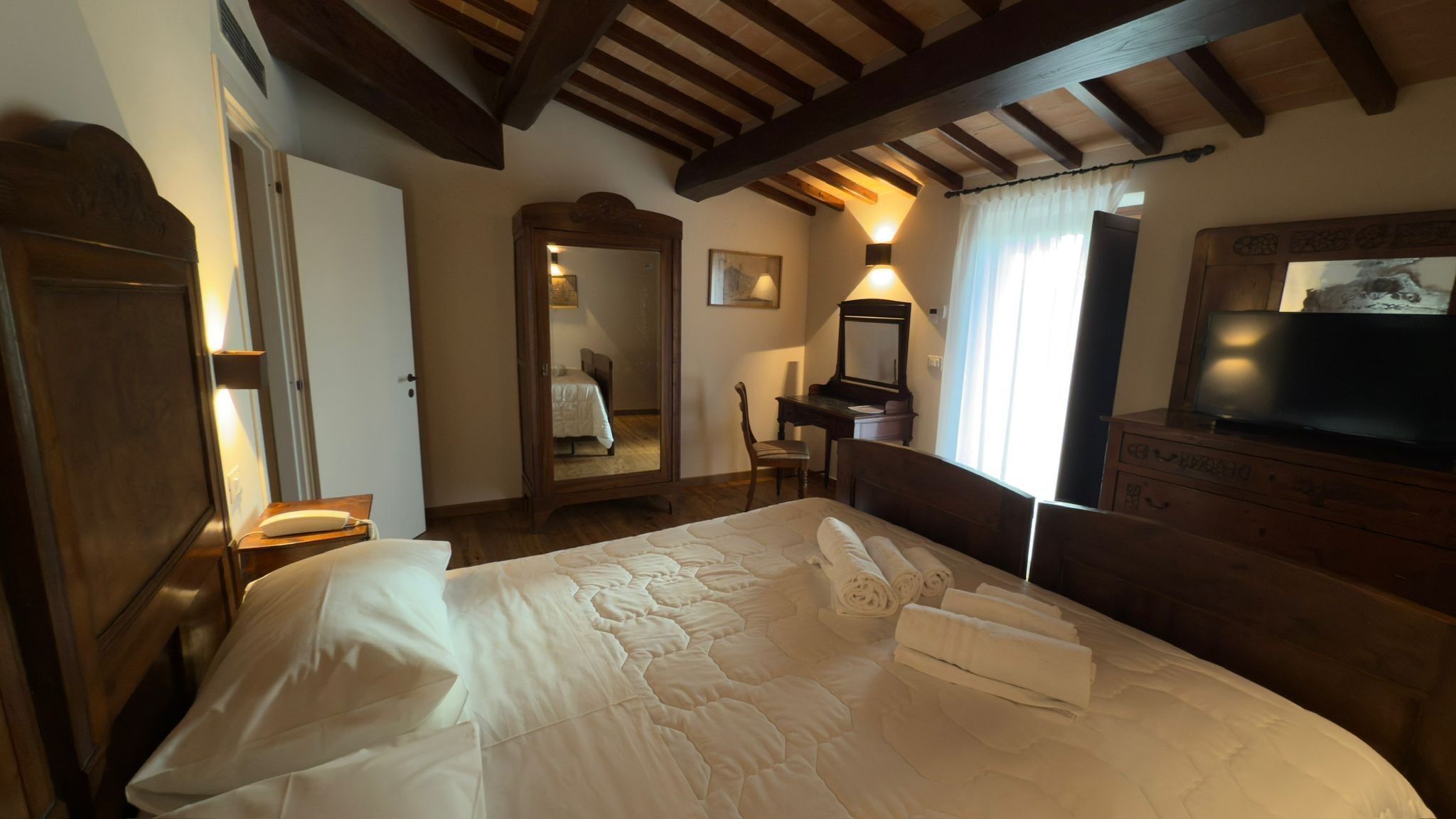 A bedroom with a large bed, a wooden wardrobe, a desk with a chair, a mirror, and a television.