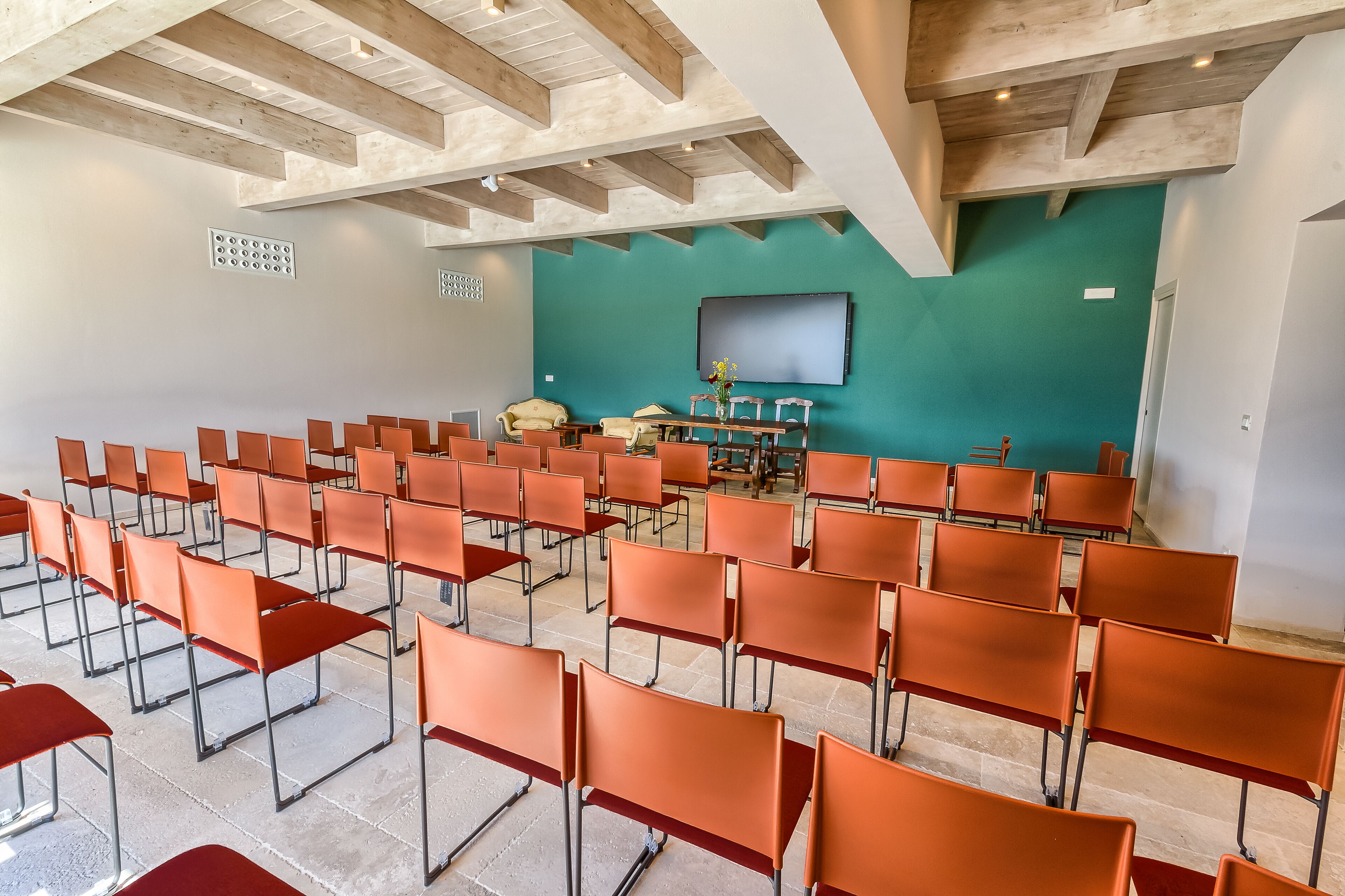 A conference room with rows of orange chairs, a teal wall, and a mounted TV.
