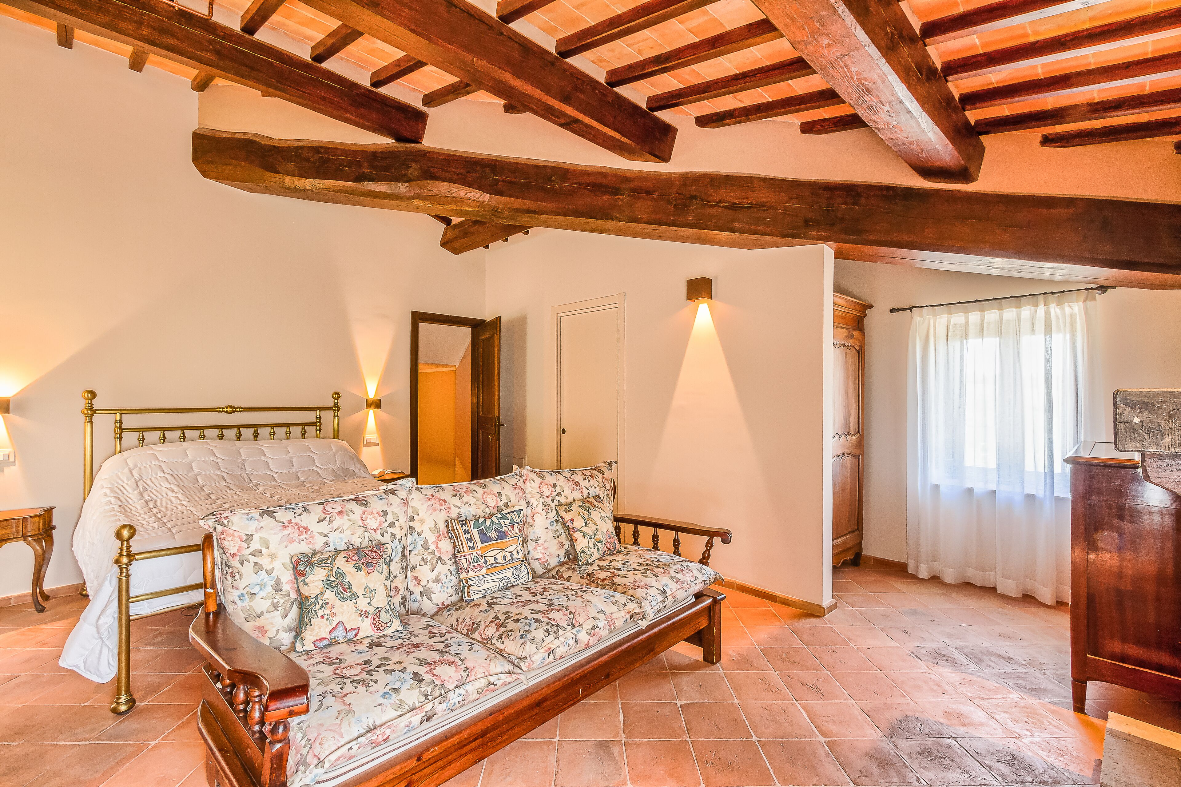 A bedroom with a bed, a wooden bench, a sofa, a wooden ceiling, a door, and a window with curtains.