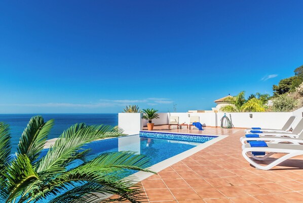 A pool area with a view of the sea, lounge chairs, and potted plants.