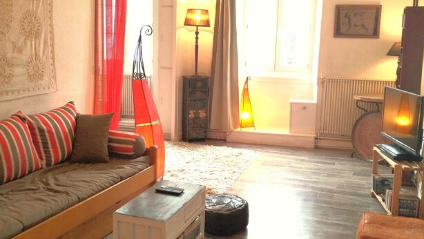 Una sala de estar acogedora con un sofá, un reposapiés y una mesita con un control remoto. Hay una lámpara, un radiador y un televisor sobre un mueble bajo.