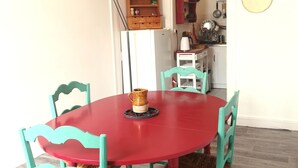 Un comedor con una mesa roja y sillas turquesas, una cafetera y una estantería con utensilios de cocina.