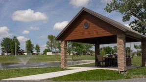 A covered pavilion with stone pillars, a seating area, and a pond in the background.