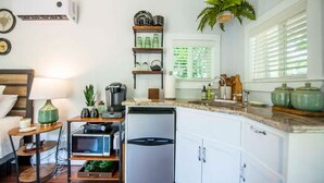 Eine kompakte Küche mit Mikrowelle, Kaffeemaschine und kleinem Kühlschrank. Es gibt ein Spülbecken mit Armatur, ein Regal mit Küchenutensilien und ein Fenster mit Jalousien.