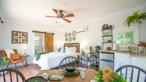 Ein gemütliches Hotelzimmer mit Bett, Esstisch mit Tellern und Schüssel, Stuhl, Deckenventilator, Küchenzeile mit Spüle und Regalen sowie einem Fenster mit Jalousien.