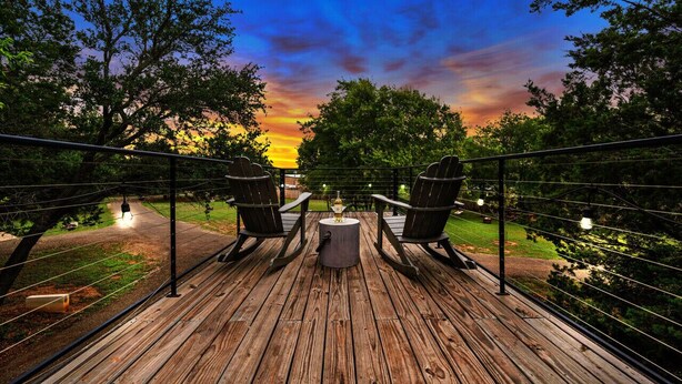 Een houten terras met twee schommelstoelen en een centrale tafel, met uitzicht op een zonsondergang en bomen op de achtergrond.