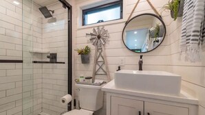 Een moderne badkamer met een glazen douchecabine, een witte wastafel met een ronde spiegel, en een decoratieve windmolen aan de muur.