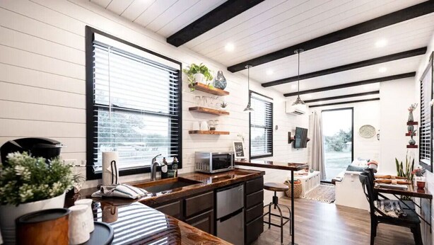 A modern kitchen with a wooden countertop, a built-in microwave, and a sink. There are shelves with plants and decorative items, and a dining area with a table and chairs.