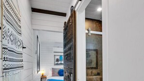 A narrow hallway with a wooden floor, a bed with a blue and white bedspread, and a wall with a patterned tapestry.