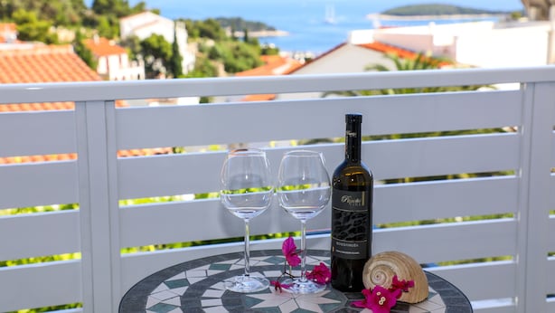 A balcony with two wine glasses, a bottle of wine, and a decorative shell on a tiled table, overlooking a coastal town. - Stella Maris Hvar - Sea View Rooms with Balcony (Hvar)