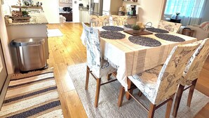 Uma sala de jantar com uma mesa posta para quatro pessoas, rodeada por cadeiras estampadas, um lustre e uma cozinha ao fundo.