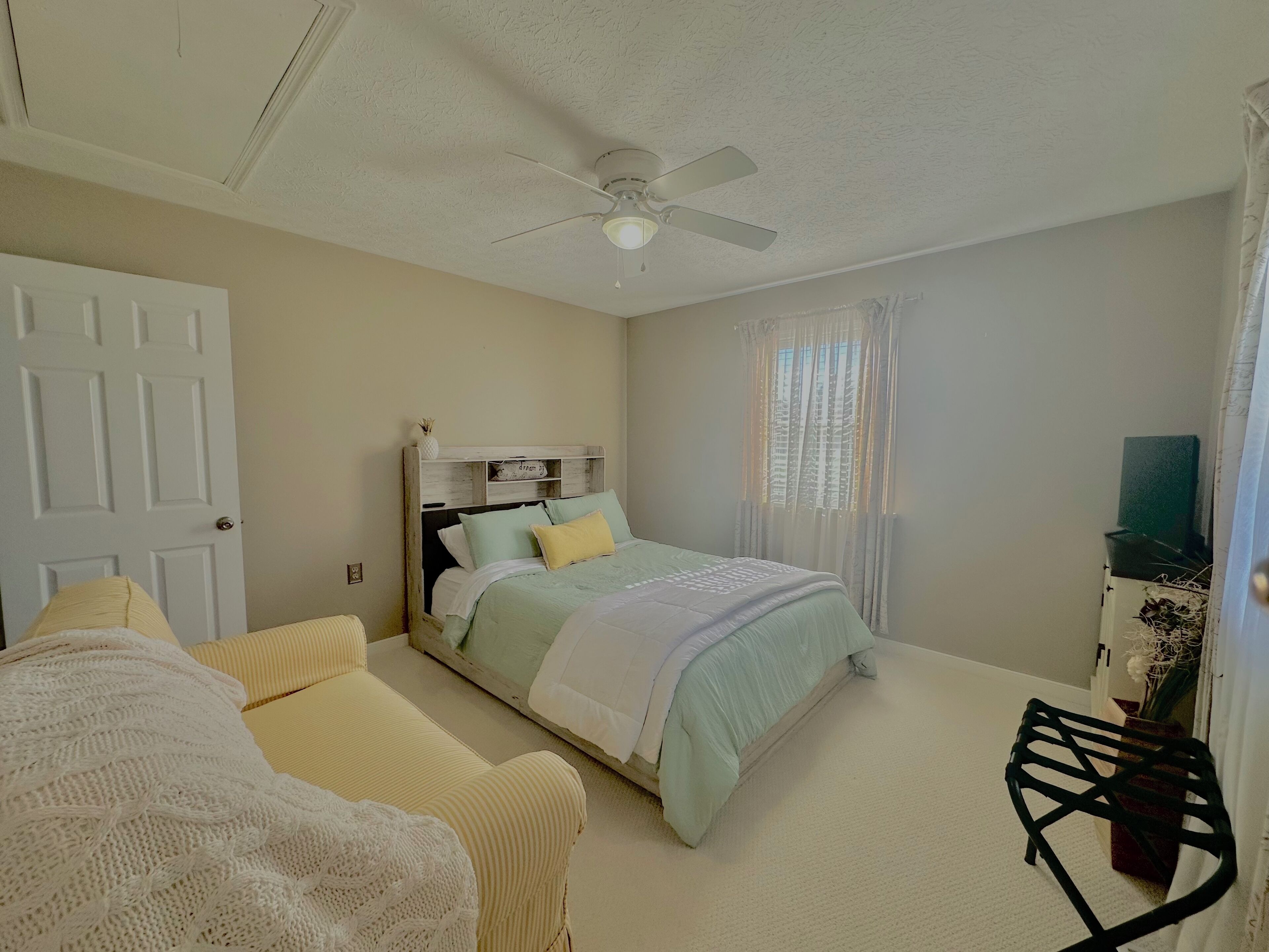 A bedroom with a bed, a chair, a ceiling fan, a door, and a window with curtains.
